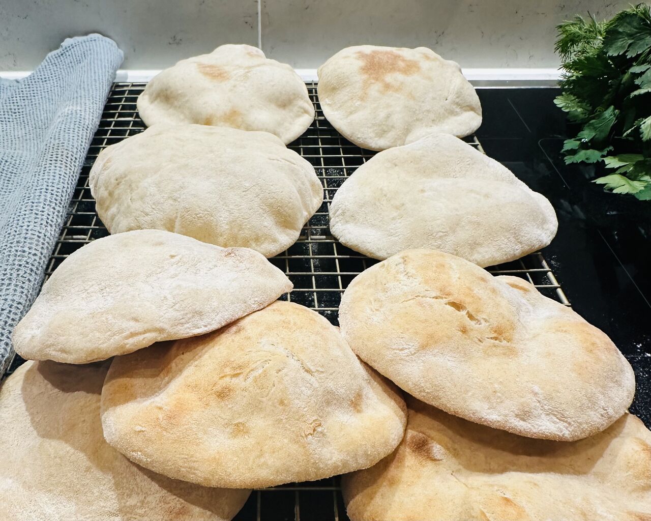 Freshly baked pita breads cooling on a black wire rack on a kitchen counter, with a light blue kitchen towel on the left and fresh parsley on the right. The pitas are puffy and golden-brown with a light dusting of flour on their surfaces.